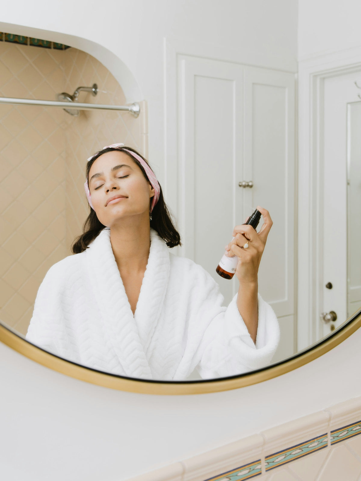 Woman in a robe misting her face at a bathroom mirror